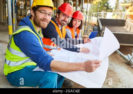 Al momento della pianificazione del lavoro, i lavoratori edili collaborano e sono architetti con la pianta del piano del cantiere Foto Stock