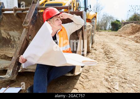 Architetto con disegno architettonico siede su una pala di escavatore e guarda il cantiere Foto Stock