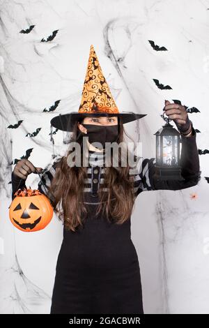 Halloween mascherato persone. Una donna in costume da strega in una maschera protettiva contiene una zucca con caramella e una lanterna luminosa. Durante la padella del coronavirus Foto Stock