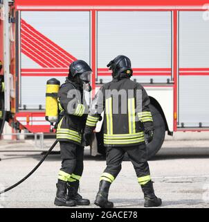 due vigili del fuoco in uniforme con bombola di ossigeno autonoma durante l'emergenza e camion antincendio in background Foto Stock