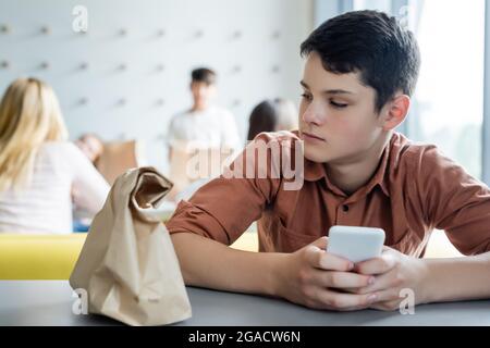 ragazzo triste con il telefono cellulare seduto da solo in un ristorante a scuola vicino borsa di carta e compagni di classe offuscati Foto Stock