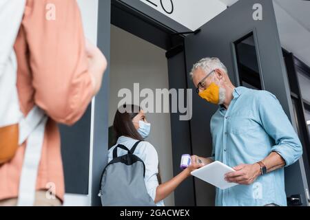 vista ad angolo basso dell'insegnante in maschera medica arancione che controlla la temperatura della studentessa con il pirometro Foto Stock