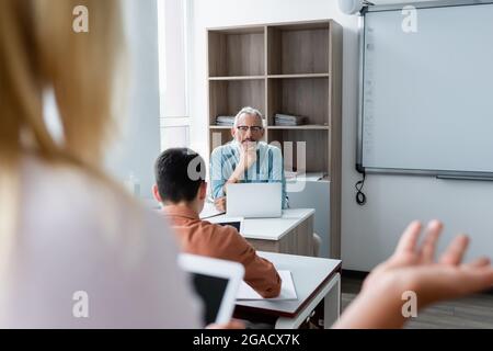 Insegnante sorridente che guarda un allievo offuscato vicino al computer portatile in classe Foto Stock