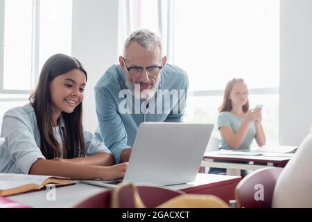 Insegnante che guarda un laptop vicino a una studentessa sorridente in classe Foto Stock