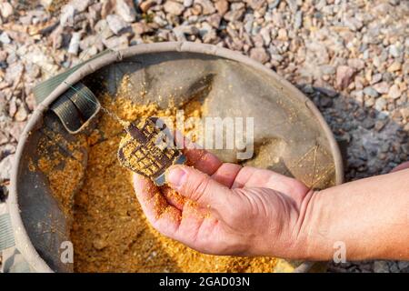 Nella mano di un pescatore un alimentatore con cibo di pesce sullo sfondo di un secchio di pesca tela in sfocatura e luce solare. Spazio di copia, primo piano. Foto Stock