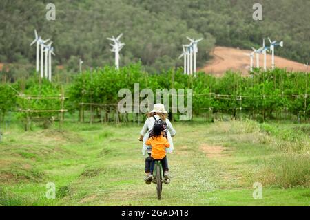 Donna e ragazzo in bicicletta in fattoria con turbine eoliche in background Foto Stock