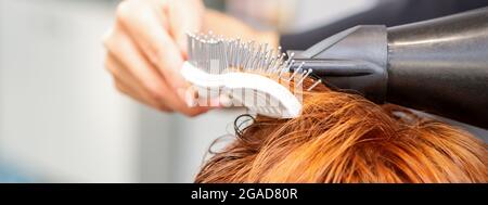 Closeup della mano del padrone con l'aria-asciugando e la spazzola che soffia i capelli rossi femminili in un salone Foto Stock