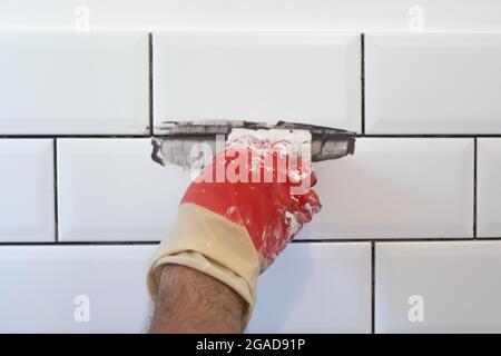 Il lavoratore applica la grota grigia a piastrelle bianche con cazzuola di gomma. Foto Stock