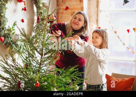 Gioiosa madre e bella figlia ragazza in felpe a maglia accogliente decorare Christmasmas abete albero con nuovo anno baubles e giocattoli in soggiorno, sorriso Foto Stock