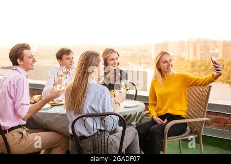 Gruppo di cinque giovani allegri che cenano al ristorante, seduti sulla terrazza a tavola e tengono in mano bicchieri di vino che posano per selfie Foto Stock