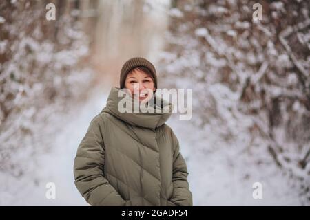 Felice giovane donna sorridente in cappello a maglia e giacca imbottita verde scuro in piedi all'aperto nel paesaggio nevoso foresta invernale dietro, guardando la fotocamera con Foto Stock