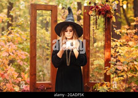 Giovane donna in costume di strega e cappello guardando misteriosamente a candela nelle sue mani isolato contro sfondo della foresta d'autunno con vintage porte di legno u Foto Stock