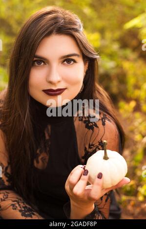 Giovane donna di brunette che tiene piccola zucca bianca di fronte al suo viso, in piedi isolato sullo sfondo della foresta d'autunno. Vacanze autunnali, Halloween dic Foto Stock