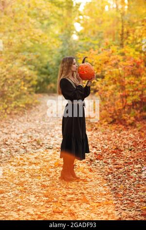 Giovane donna bionda che tiene spaventosa zucca artigianale di fronte al viso, in piedi isolato sullo sfondo della foresta autunnale. Vacanze autunnali, Halloween deco Foto Stock