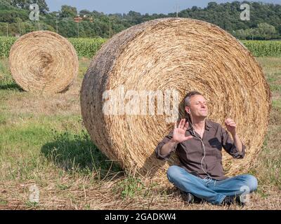 Un uomo bianco di mezza età ascolta la musica sulle cuffie seduto in un campo, con la schiena a una balla di fieno Foto Stock