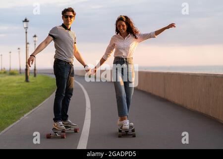 Coppia di skateboarder ride longboard che tiene le mani. I giovani adulti felici godono di longboard al tramonto Foto Stock