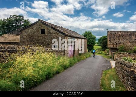 Ciclisti in bicicletta lungo una strada di campagna tra vecchi edifici di pietra fattoria, Stainburn, North Yorkshire, Regno Unito Foto Stock