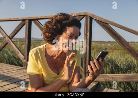 Una donna allegra e vitale, nei suoi anni '70, si saluta mentre chiacchiera con qualcuno in videochiamata da una zona naturale del Mediterraneo, Spagna Foto Stock