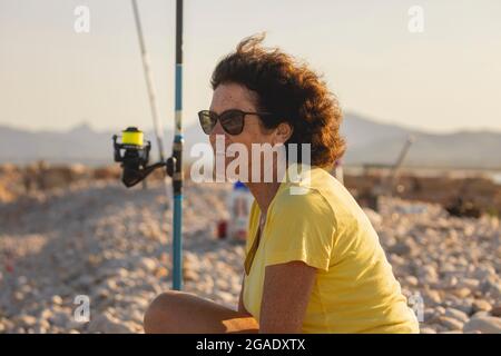 Una donna allegra e vitale, negli anni '70, che pesca con la canna da pesca, su una spiaggia naturale, al tramonto, accanto al Mar Mediterraneo, Spagna Foto Stock