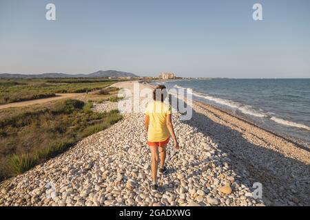 Una donna allegra e vitale, nei suoi anni '70, cammina lungo una spiaggia di ghiaia naturale al tramonto, vicino al Mar Mediterraneo, Spagna Foto Stock