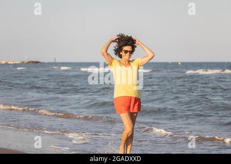 Una donna allegra e vitale, nei suoi anni '70, cammina su una spiaggia al tramonto, sulle rive del Mar Mediterraneo, Spagna Foto Stock