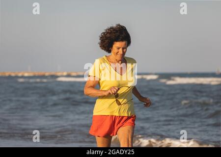 Una donna allegra e vitale, nei suoi anni '70, cammina e corre su una spiaggia al tramonto, sulle rive del Mar Mediterraneo, Spagna Foto Stock
