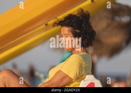 Una donna adulta, negli anni 70, seduta tranquilla sulla spiaggia, pensando e meditando, mentre prendisole, Mediterraneo, Spagna Foto Stock