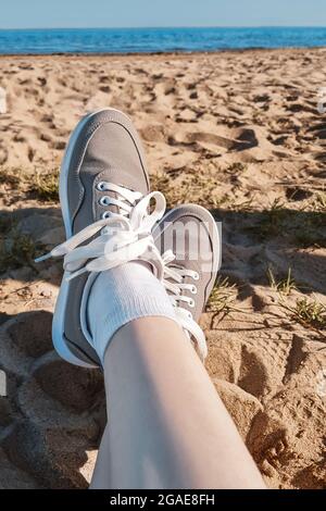 Gambe in sneaker grigie sulla spiaggia di sabbia. Concetto di vacanza estiva vicino al mare. Viaggi lifestyle. Vista POV. Spazio di copia. Foto Stock