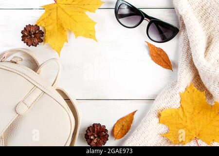 Ciao autunno. Composizione creativa piatta su sfondo bianco. Foglie gialle, maglione a maglia, zaino e occhiali. Spazio di copia. Foto Stock
