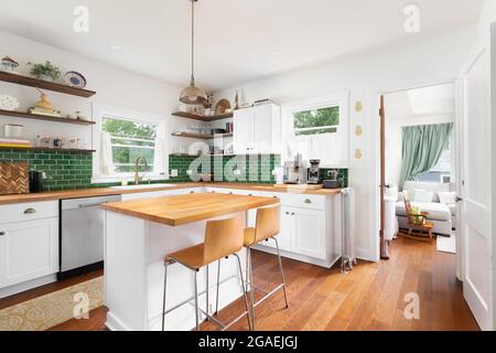 Una cucina rinnovata con armadietti bianchi, un piano di lavoro in legno naturale, sedie che si trovano sull'isola, e un backsplash di piastrelle verdi della metropolitana. Foto Stock