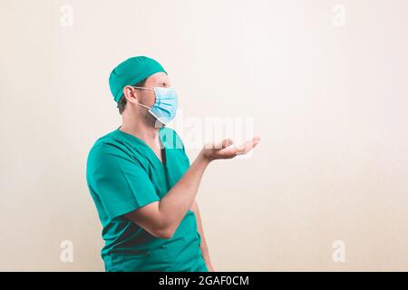 Giovane medico ritratto in tuta verde chirurgo e maschera protettiva di respirazione con nulla in mano sullo sfondo giallo isolato Foto Stock