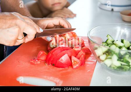 Donna anziana nonna cucina insalata sana per nipote. Nonna che taglia il pomodoro con un coltello sul tagliere per la cena Foto Stock