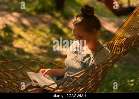 Giovane studentessa sorridente a leggere un libro mentre si steso su un'amaca in giardino. Donna che si rilassa nel libro di lettura amaca. Torna all'università, all'università. Foto Stock