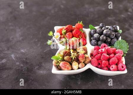Una miscela di bacche fresche in un piatto bianco su sfondo grigio scuro, alimentazione naturale cruda Foto Stock