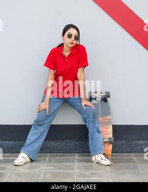 Giovane donna asiatica in T-shirt rossa con occhiali da sole riposati dopo lo skateboard. Il suo skateboard si appoggia contro un muro sul marciapiede. Foto Stock