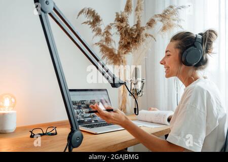 Giovane donna allegra che registra podcast in casa studio. Podcaster femmina che registra la trasmissione nel microfono Foto Stock