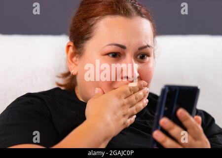 Primo piano foto di una donna di capelli rossi di grandi dimensioni in nero t-shirt guardare cattive notizie utilizzando Internet mobile e lei sembra molto consapevolezza, coprendola Foto Stock
