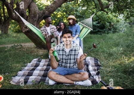 Bel ragazzo indiano bere birra e mangiare salsicce alla griglia durante il picnic sulla natura. Tre amici diversi che riposano in amaca sullo sfondo. Concetto di estate. Foto Stock