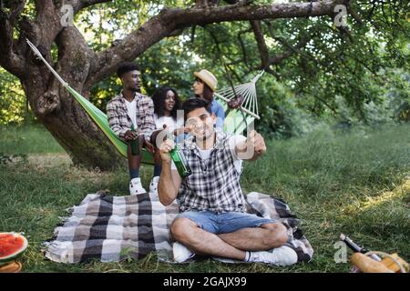 Giovane uomo in stile casual seduto su coperta a scacchi con salsicce alla griglia e bottiglia di birra in mano. Sfondo sfocato di amici multirazziali che si aggirano in amaca. Picnic nel giardino estivo. Foto Stock