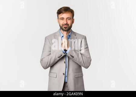 Ritratto di bell'uomo bearded in abito grigio, ringraziando per qualcosa, pregando di aiuto, sorridendo speranzoso, in piedi su sfondo bianco Foto Stock