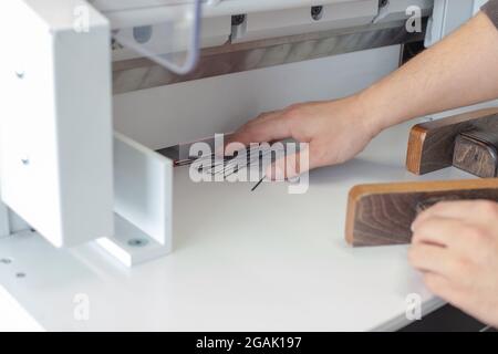 un operatore specializzato rimuove i frammenti e i bordi di una pila di biglietti da visita o etichette su una macchina automatica per il taglio della carta, una taglierina professionale Foto Stock