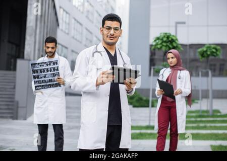 Tre giovani medici che indossano camice bianco, che lavorano con tablet pc, raggi X e cartella clinica del paziente fuori dall'ospedale. Medico indù maschile digitando sul computer tablet, in piedi di fronte ai colleghi Foto Stock