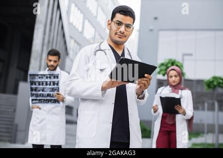 Tre giovani medici che indossano camice bianco, che lavorano con tablet pc, raggi X e cartella clinica del paziente fuori dall'ospedale. Medico indù maschile digitando sul computer tablet, in piedi di fronte ai colleghi Foto Stock