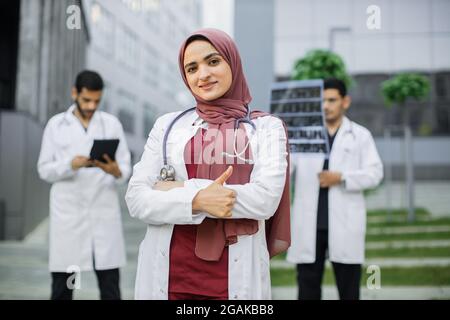 Bella giovane dottoressa araba in uniforme e hijab sta guardando la macchina fotografica e sorridendo con il pollice in su mentre i suoi due colleghi di fiducia maschile sono in piedi in background all'aperto vicino all'ospedale moderno Foto Stock