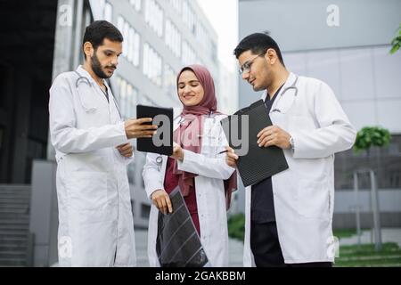 Tre giovani medici multirazziali fiduciosi in uniforme medica, donna in hijab e due uomini, guardando TAC scansione x-ray su tablet pc. L'équipe medica araba ha una discussione all'aperto di fronte alla clinica Foto Stock