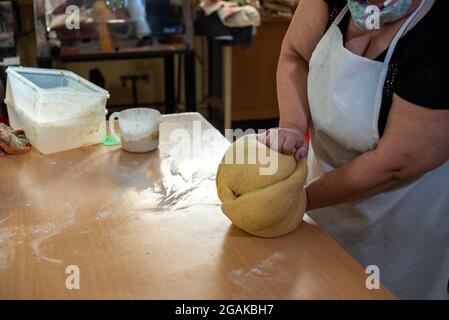 Mujer caucásica masando masa para hacer pan y bizcochos en una mesa color madera con luz de día, en una panadería. Foto Stock