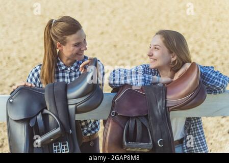 Due giovani ragazze in piedi con le mani poggiate sulla recinzione di legno nel ranch di cavalli. Ragazze di buon umore che ridono e parlano. Selle in pelle appese sulla recinzione in legno in primo piano. Foto Stock