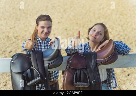 Due ragazze giovani in piedi dietro la recinzione di legno nel ranch di cavalli. Ragazze che posano per la macchina fotografica e fanno un segno di pollice-su. Selle in pelle appese sulla recinzione in legno in primo piano. Foto Stock