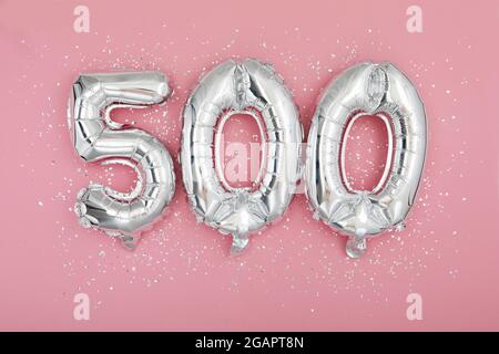 Dall'alto di palloncini argentati brillanti che mostrano il numero 500 su sfondo rosa con glitter sparso Foto Stock
