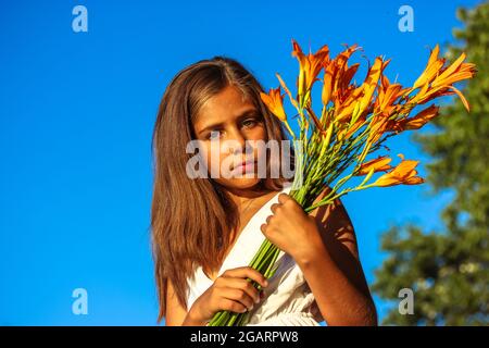 Una bella ragazza adolescente seria in abito bianco con un bouquet di gigli d'arancio contro un cielo blu in estate soleggiato giorno. Capretto femmina di 10-12 anni. Foto Stock